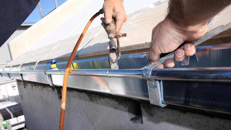 Arbeiter befestigt ein Metallteil mit einer Zange an einer Dachrinne, während ein Schlauch im Hintergrund sichtbar ist.