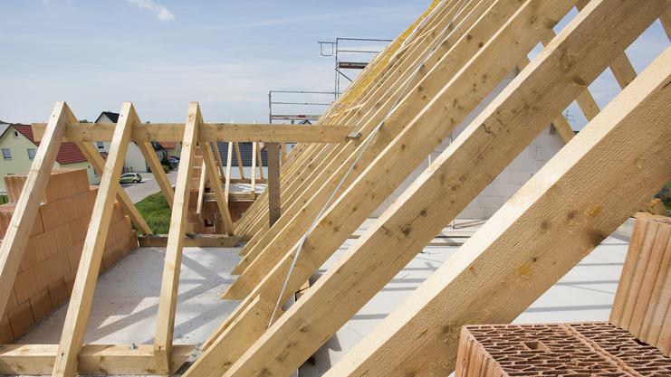 Blick auf eine Dachkonstruktion mit Holzsparren und Ziegelsteinen auf einer Baustelle unter blauem Himmel.