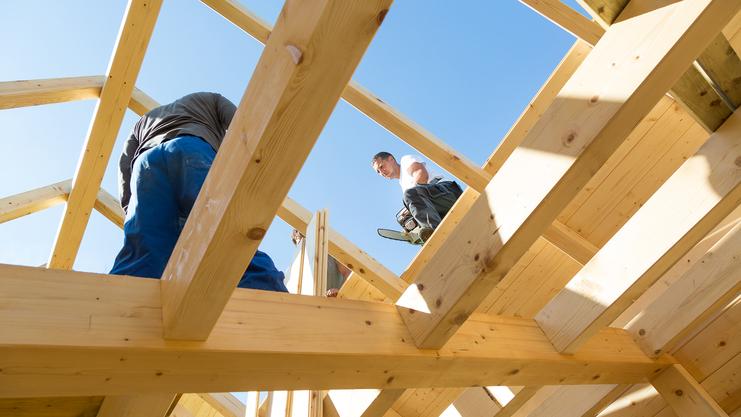 Blick von unten auf zwei Handwerker, die an einem Holzgerüst unter blauem Himmel arbeiten.